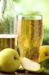 Photo of Delicious cider in glass, bottle, apples and green leaves on table outdoors, closeup