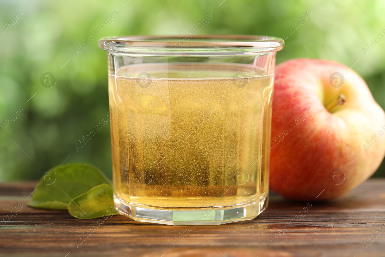 Photo of Delicious cider in glass, apple and green leaves on wooden table outdoors, closeup