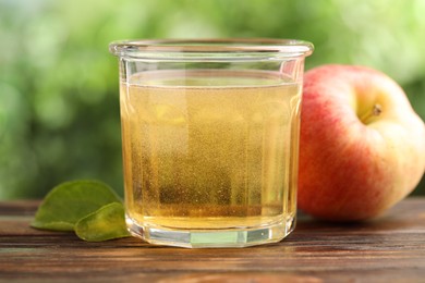 Photo of Delicious cider in glass, apple and green leaves on wooden table outdoors, closeup