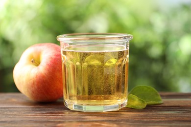 Photo of Delicious cider in glass, apple and green leaves on wooden table outdoors, closeup