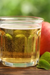 Photo of Delicious cider in glass, apple and green leaves on wooden table outdoors, closeup