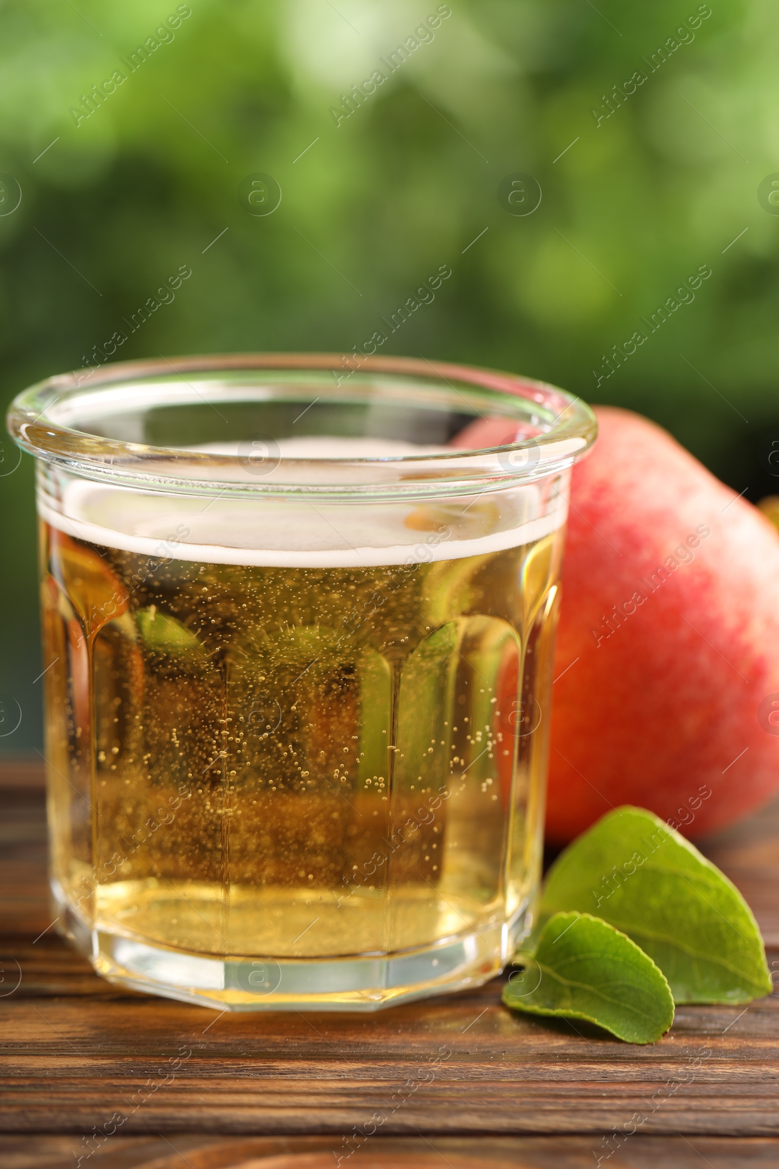 Photo of Delicious cider in glass, apple and green leaves on wooden table outdoors, closeup