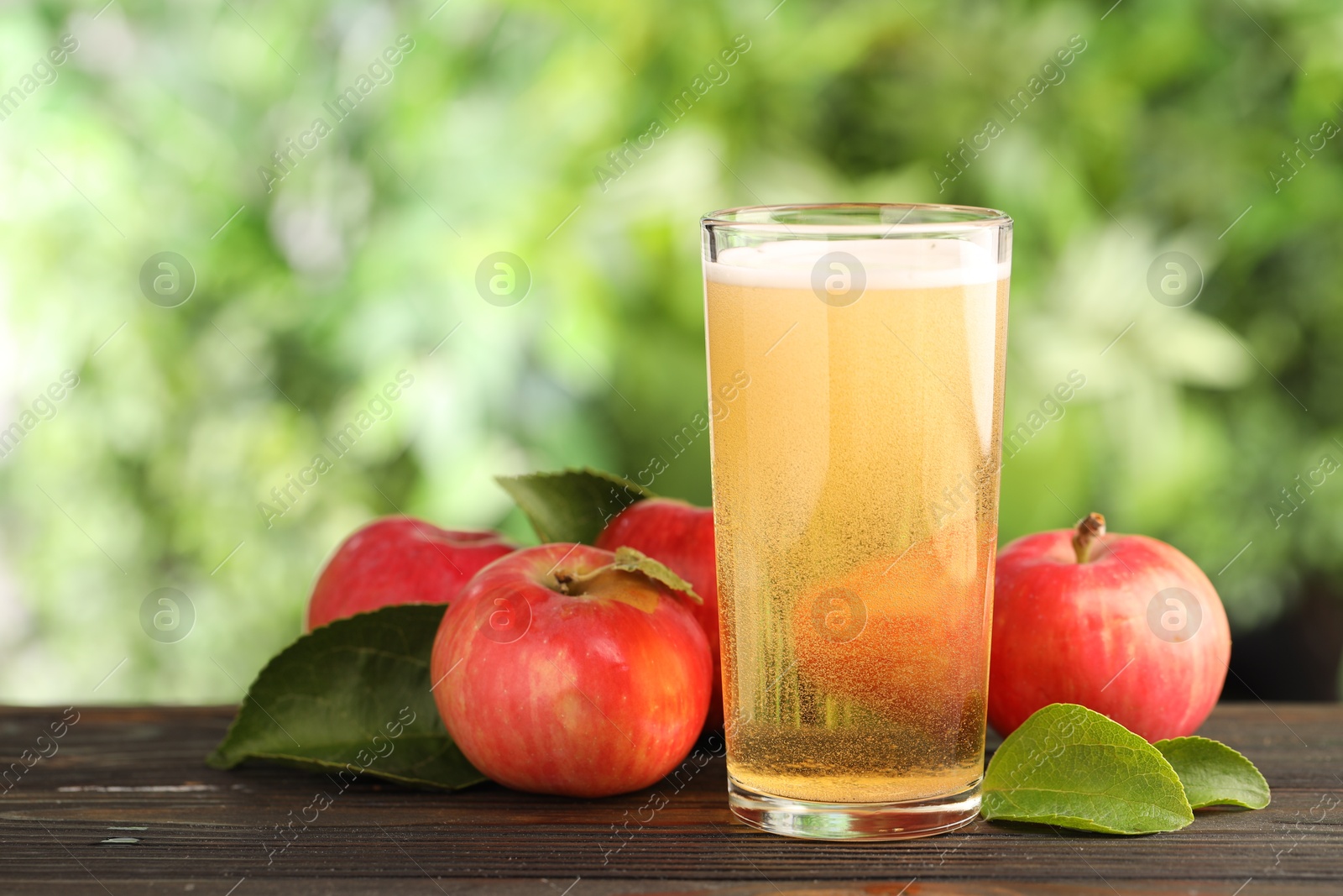 Photo of Delicious cider in glass, apples and green leaves on wooden table outdoors