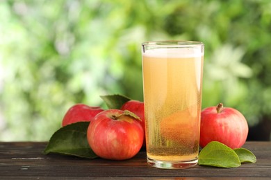 Photo of Delicious cider in glass, apples and green leaves on wooden table outdoors