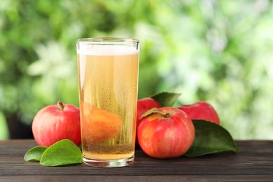Photo of Delicious cider in glass, apples and green leaves on wooden table outdoors