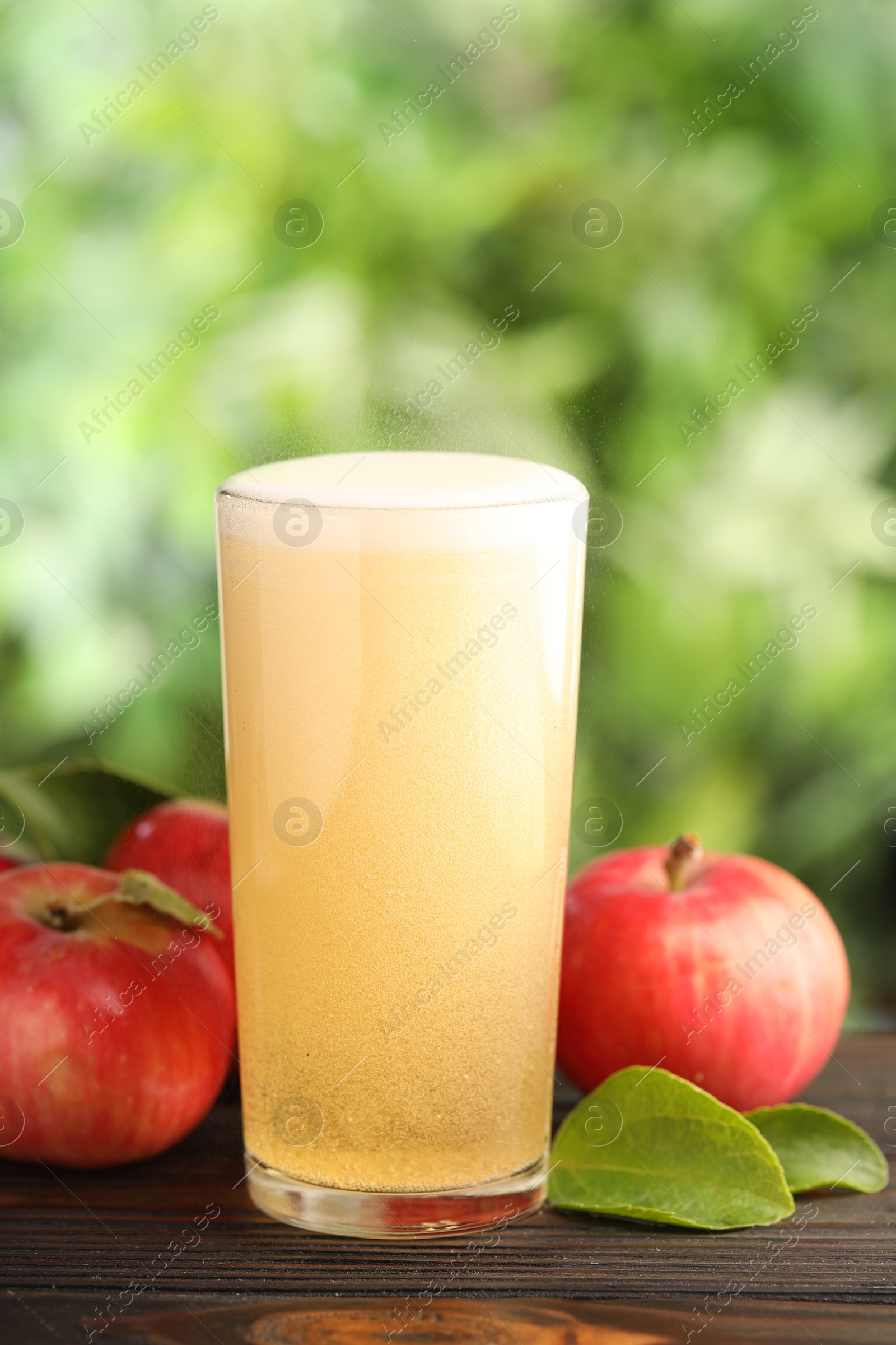 Delicious cider in glass, apples and green leaves on wooden table outdoors, closeup Photo of Delicious cider in glass, apples and green leaves on wooden table outdoors, closeup