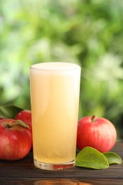 Photo of Delicious cider in glass, apples and green leaves on wooden table outdoors, closeup