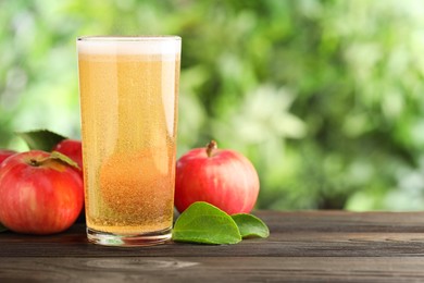 Photo of Delicious cider in glass, apples and green leaves on wooden table outdoors, space for text