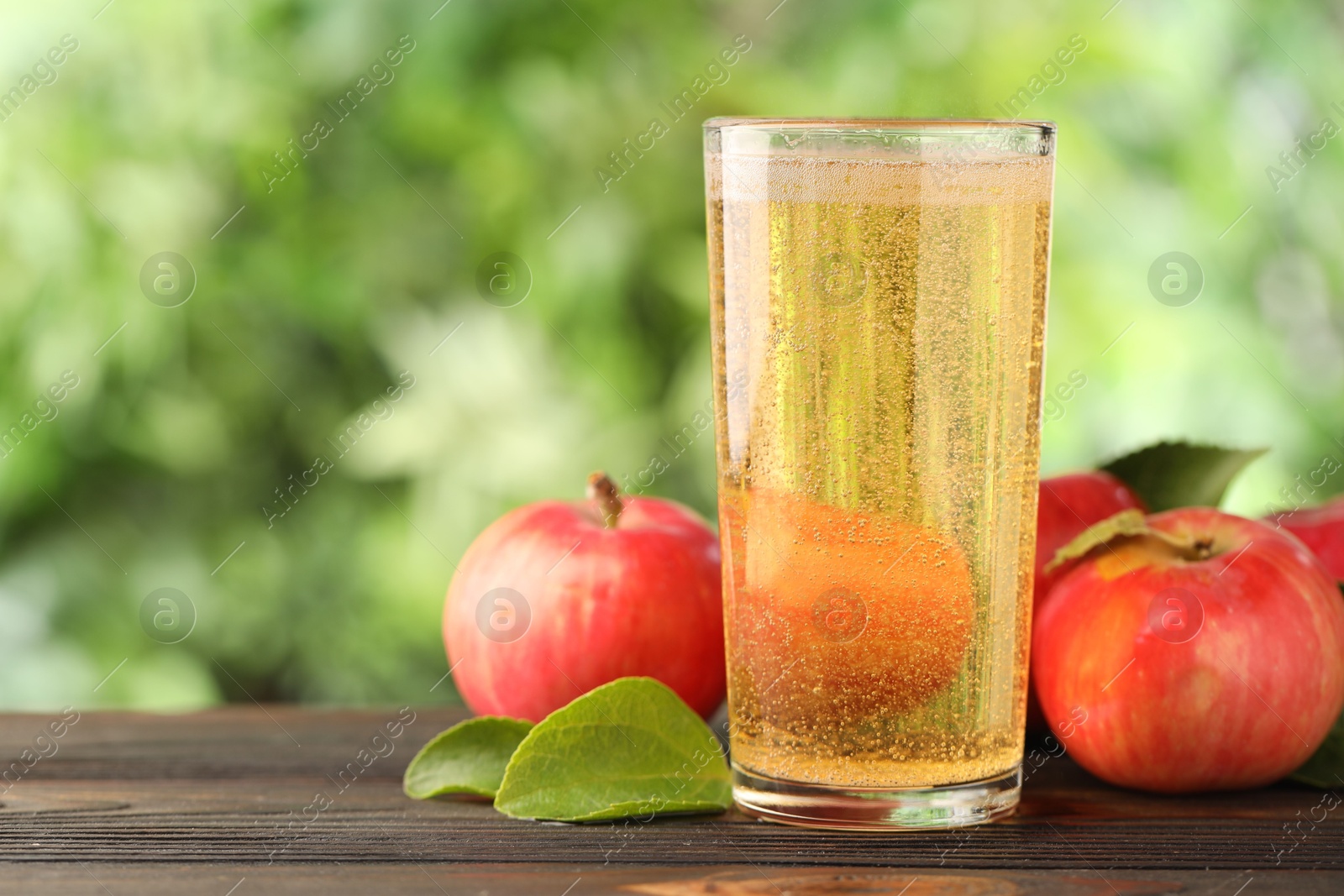 Delicious cider in glass, apples and green leaves on wooden table outdoors, space for text Photo of Delicious cider in glass, apples and green leaves on wooden table outdoors, space for text