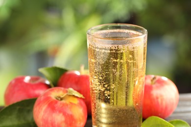 Photo of Delicious cider in glass, apples and green leaves on table outdoors, closeup
