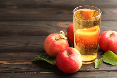 Photo of Delicious cider in glass, apples and green leaves on wooden table, closeup. Space for text