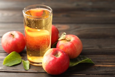 Photo of Delicious cider in glass, apples and green leaves on wooden table, closeup. Space for text
