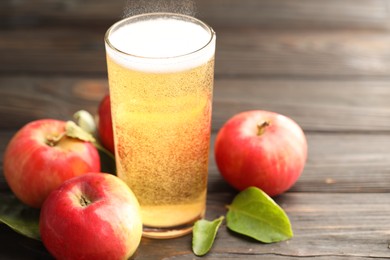 Photo of Delicious cider in glass, apples and green leaves on wooden table, closeup