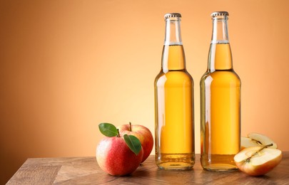 Photo of Delicious cider in glass bottles and apples on wooden table against orange gradient background, space for text