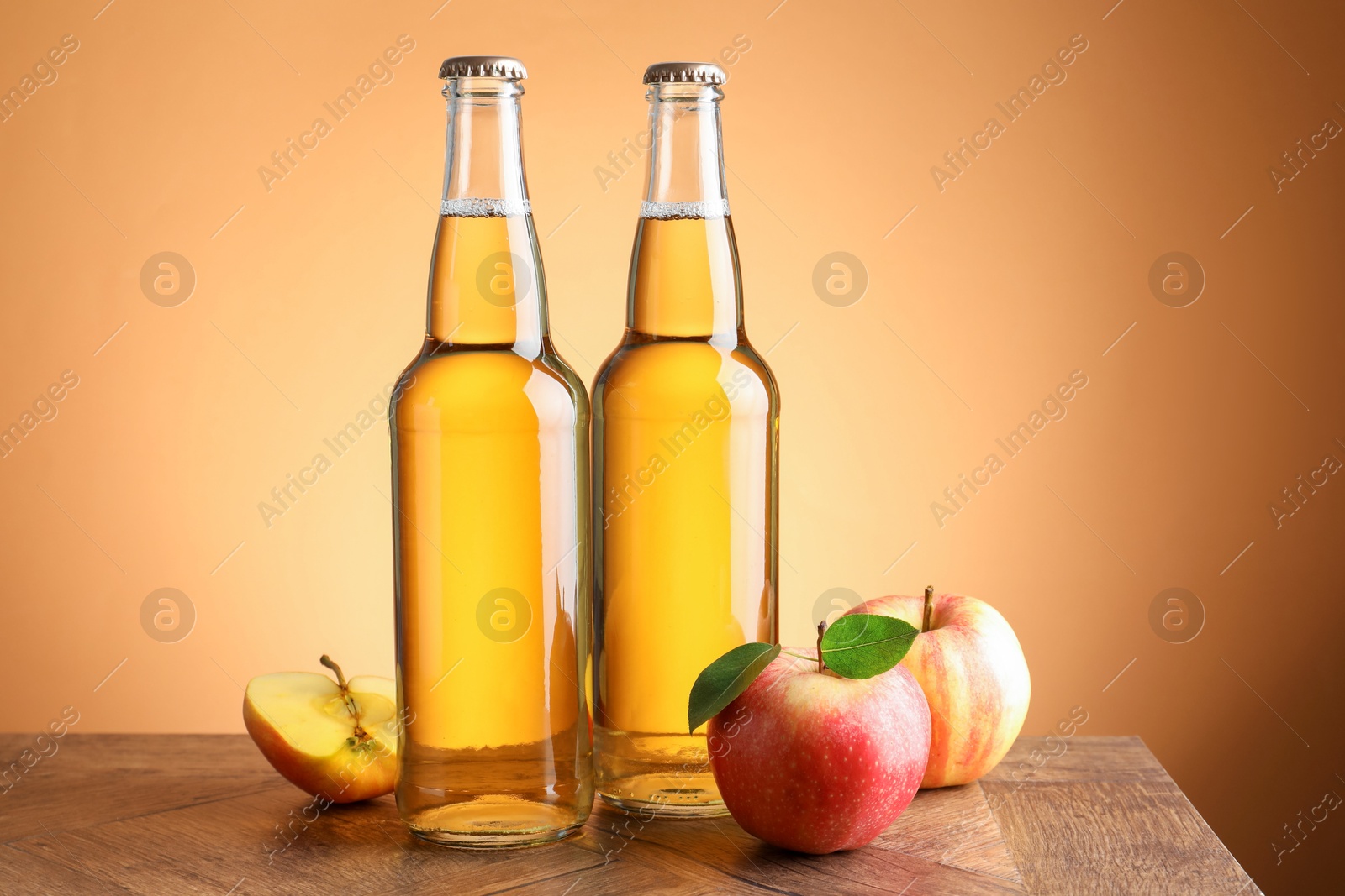 Photo of Delicious cider in glass bottles and apples on wooden table against orange gradient background