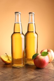 Photo of Delicious cider in glass bottles and apples on wooden table against orange gradient background