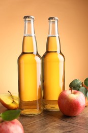 Photo of Delicious cider in glass bottles and apples on wooden table against orange gradient background
