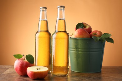 Photo of Delicious cider in glass bottles and apples on wooden table against orange gradient background