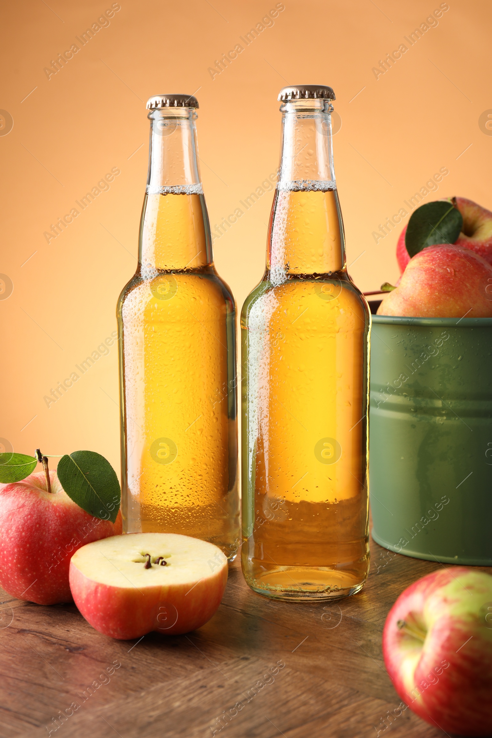 Photo of Delicious cider in glass bottles and apples on wooden table against orange gradient background