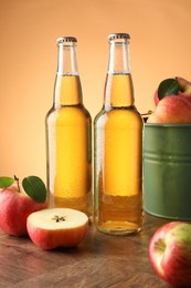 Photo of Delicious cider in glass bottles and apples on wooden table against orange gradient background