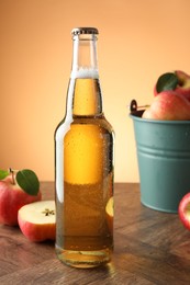 Photo of Delicious cider in glass bottle and apples on wooden table against orange gradient background