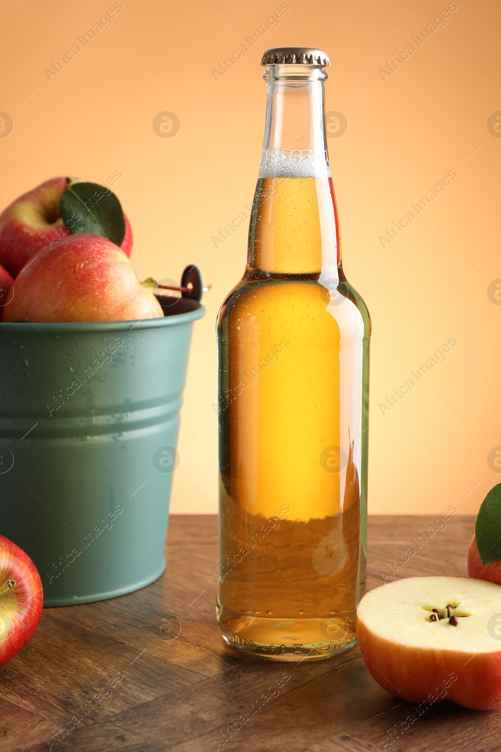 Photo of Delicious cider in glass bottle and apples on wooden table against orange gradient background