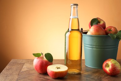 Photo of Delicious cider in glass bottle and apples on wooden table against orange gradient background. Space for text