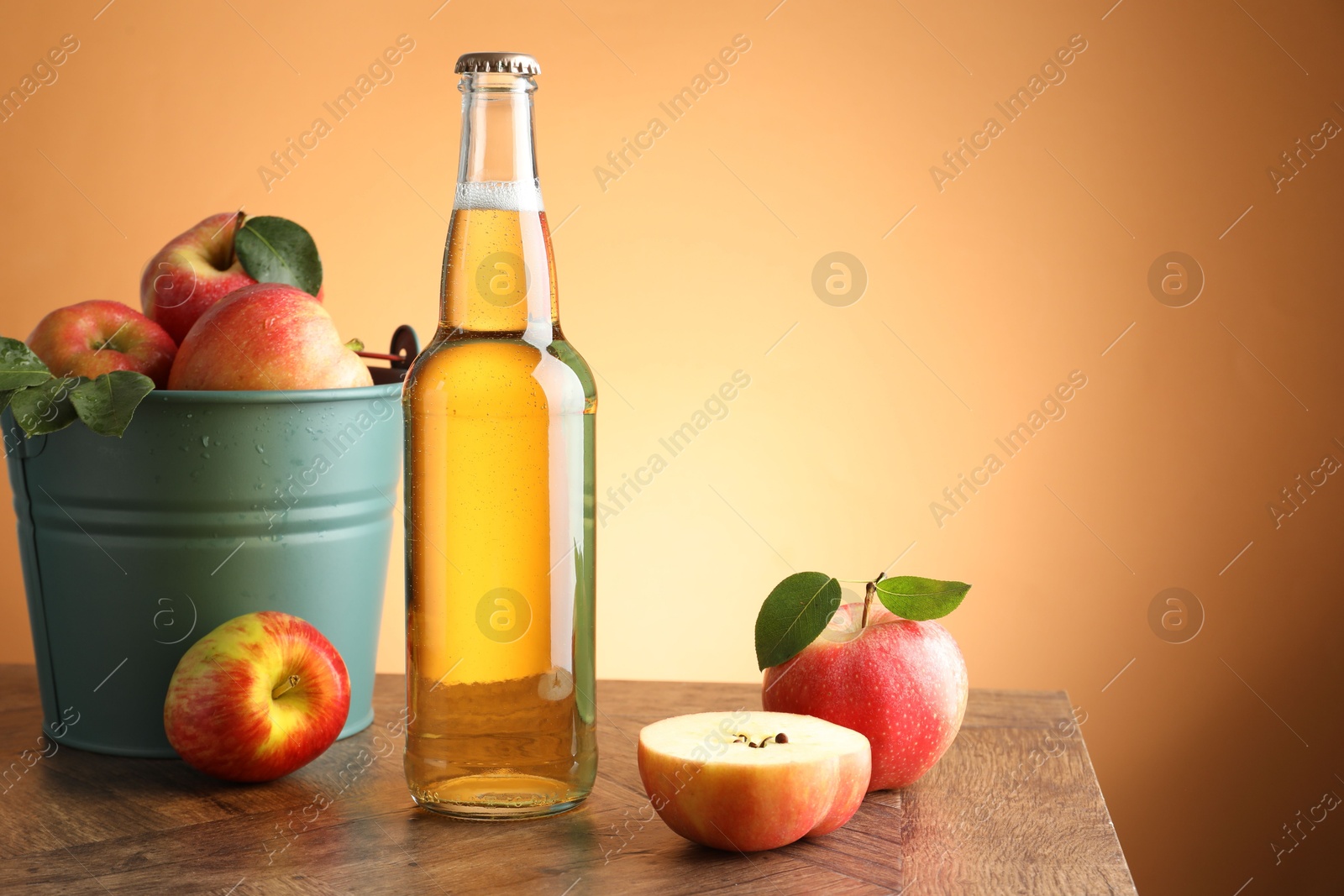 Photo of Delicious cider in glass bottle and apples on wooden table against orange gradient background. Space for text