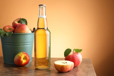Photo of Delicious cider in glass bottle and apples on wooden table against orange gradient background. Space for text