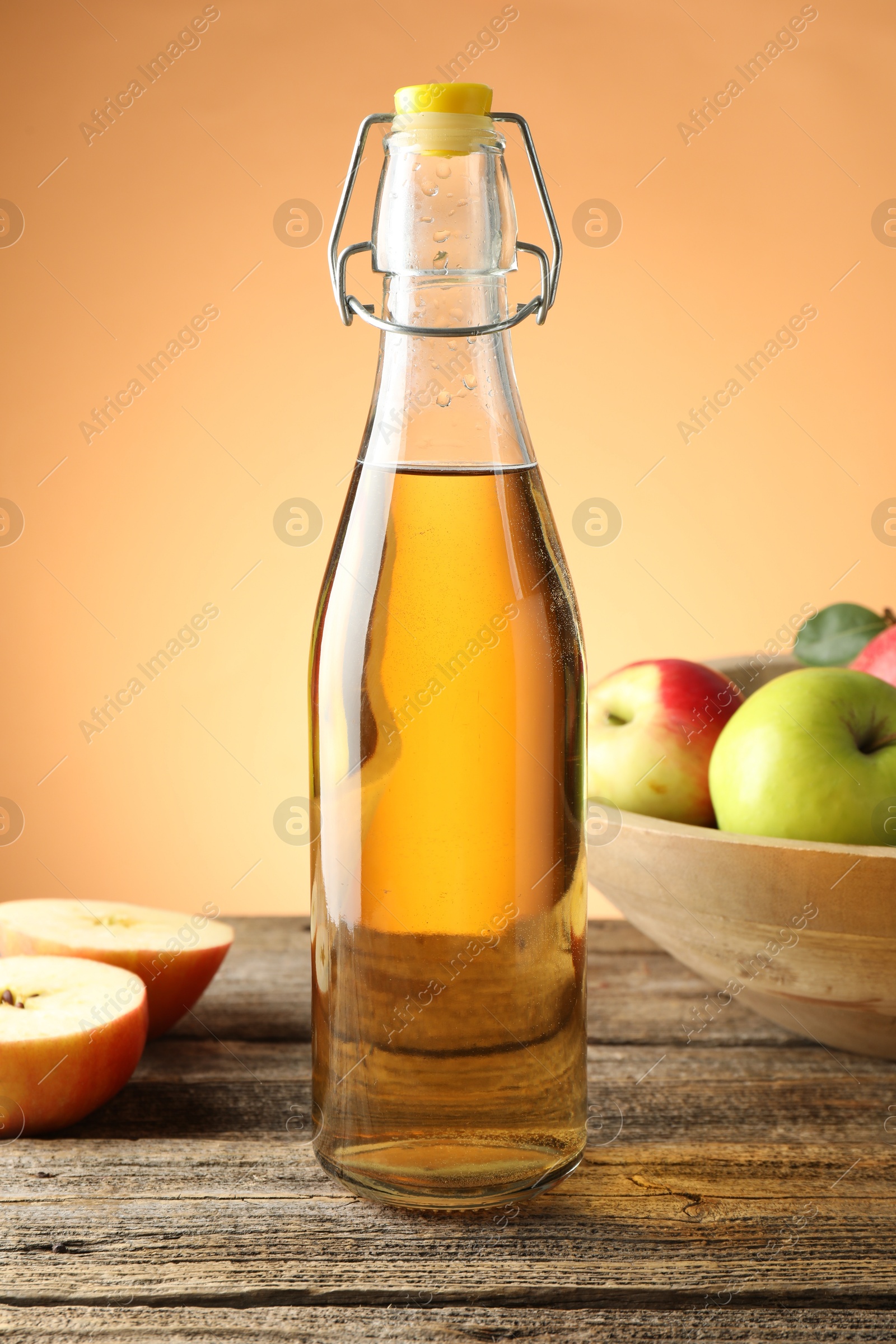 Photo of Delicious cider in glass bottle and apples on wooden table against orange gradient background