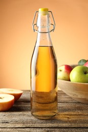Photo of Delicious cider in glass bottle and apples on wooden table against orange gradient background