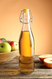 Photo of Delicious cider in glass bottle and apples on wooden table against orange gradient background