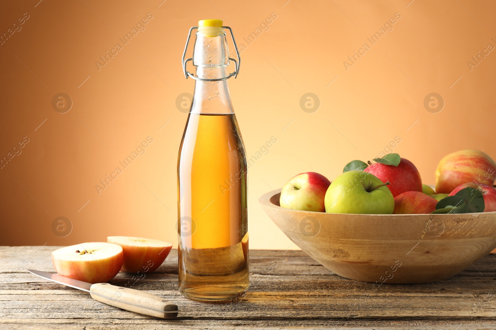 Photo of Delicious cider in glass bottle, apples and knife on wooden table against orange gradient background