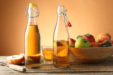 Photo of Delicious cider in bottles, glass, apples and knife on wooden table against orange gradient background