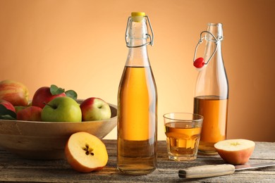 Photo of Delicious cider in bottles, glass, apples and knife on wooden table against orange gradient background