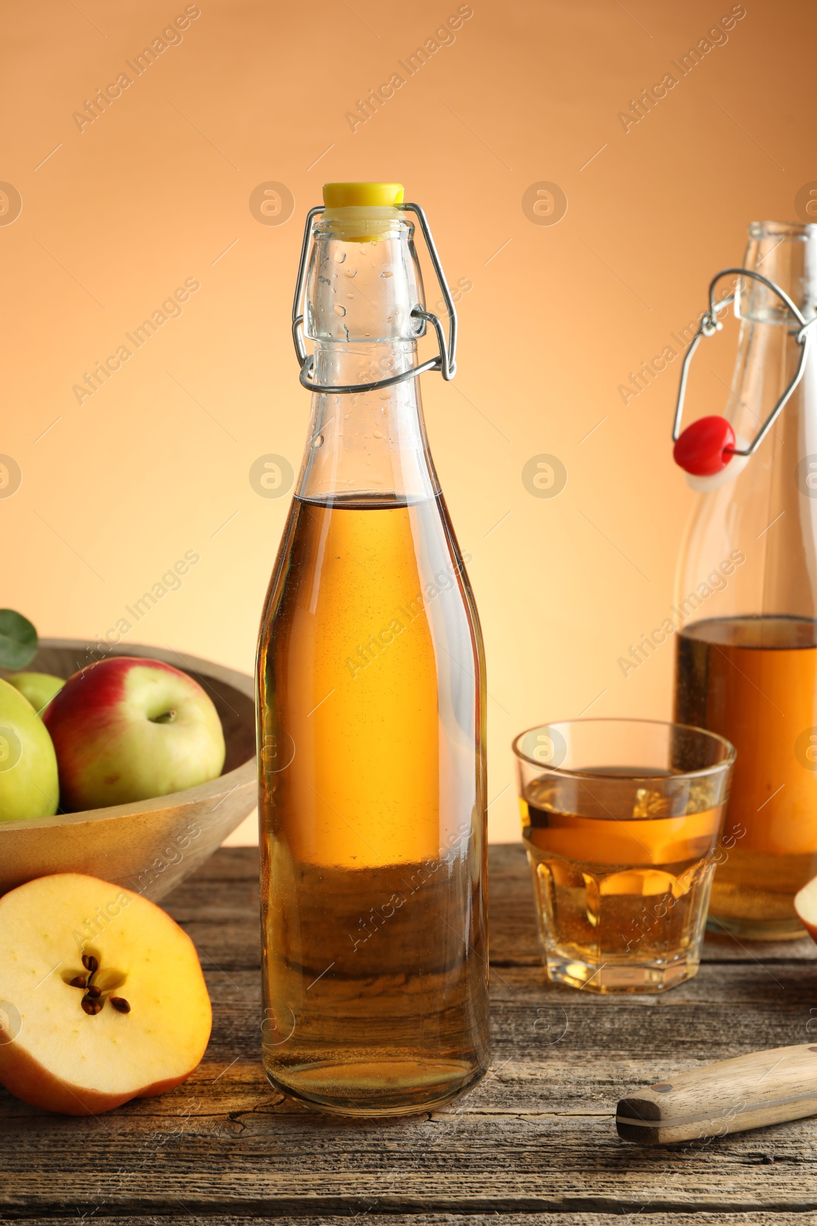 Photo of Delicious cider in bottles, glass and apples on wooden table against orange gradient background, closeup
