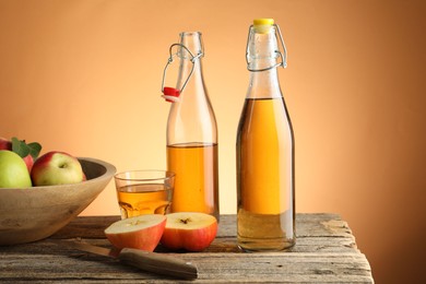 Photo of Delicious cider in bottles, glass, apples and knife on wooden table against orange gradient background