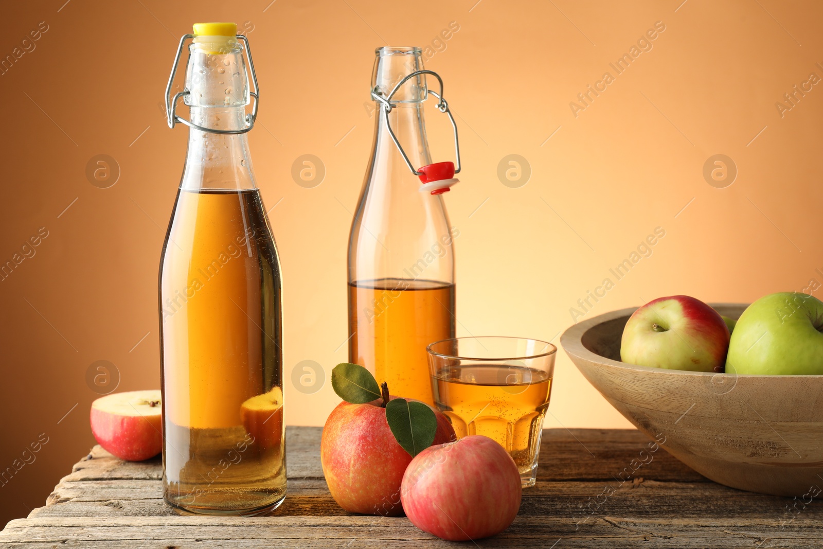 Photo of Delicious cider in bottles, glass and apples on wooden table against orange gradient background
