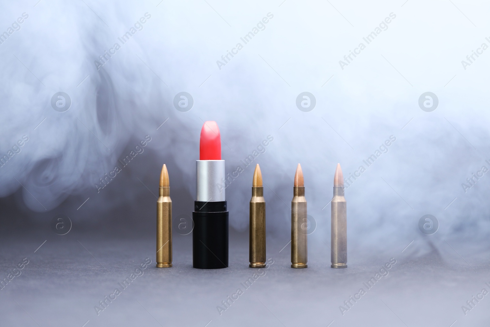 Red lipstick among bullets in smoke on grey background Photo of Red lipstick among bullets in smoke on grey background