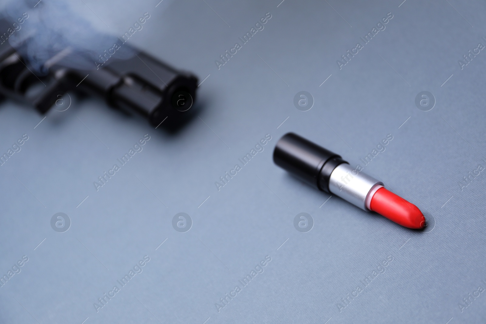 Photo of Gun and red lipstick as bullet in smoke on grey background, closeup