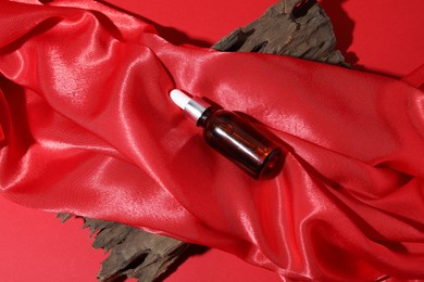 Photo of Promotion. Bottle of cosmetic product, red silk fabric and driftwood on color background, top view