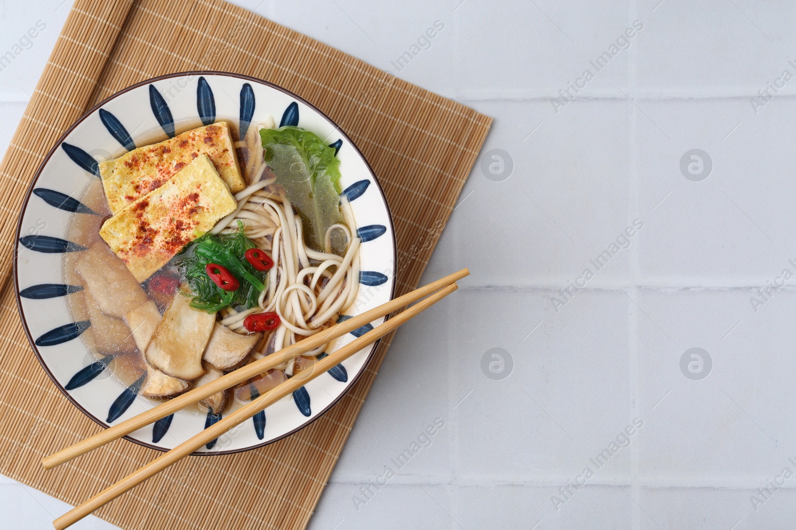 Photo of Tasty ramen with king oyster (eryngii) mushrooms in bowl on white tiled table, top view. Space for text