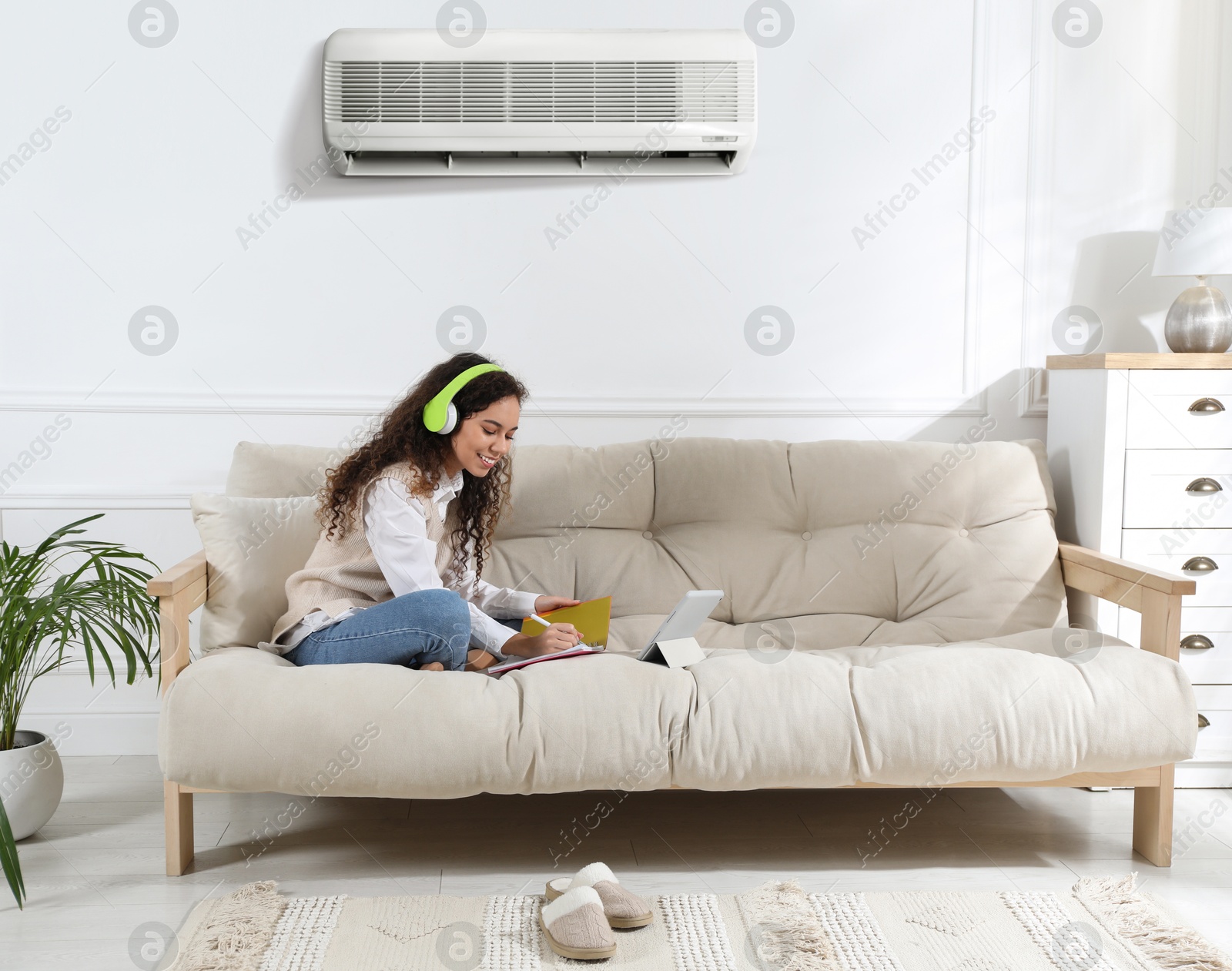 Image of Woman with headphones and tablet studying on sofa at home. Air conditioner keeping comfortable temperature in room