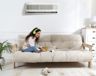 Image of Woman with headphones and tablet studying on sofa at home. Air conditioner keeping comfortable temperature in room