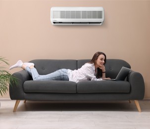 Image of Smiling woman with laptop lying on sofa at home. Air conditioner keeping comfortable temperature in room