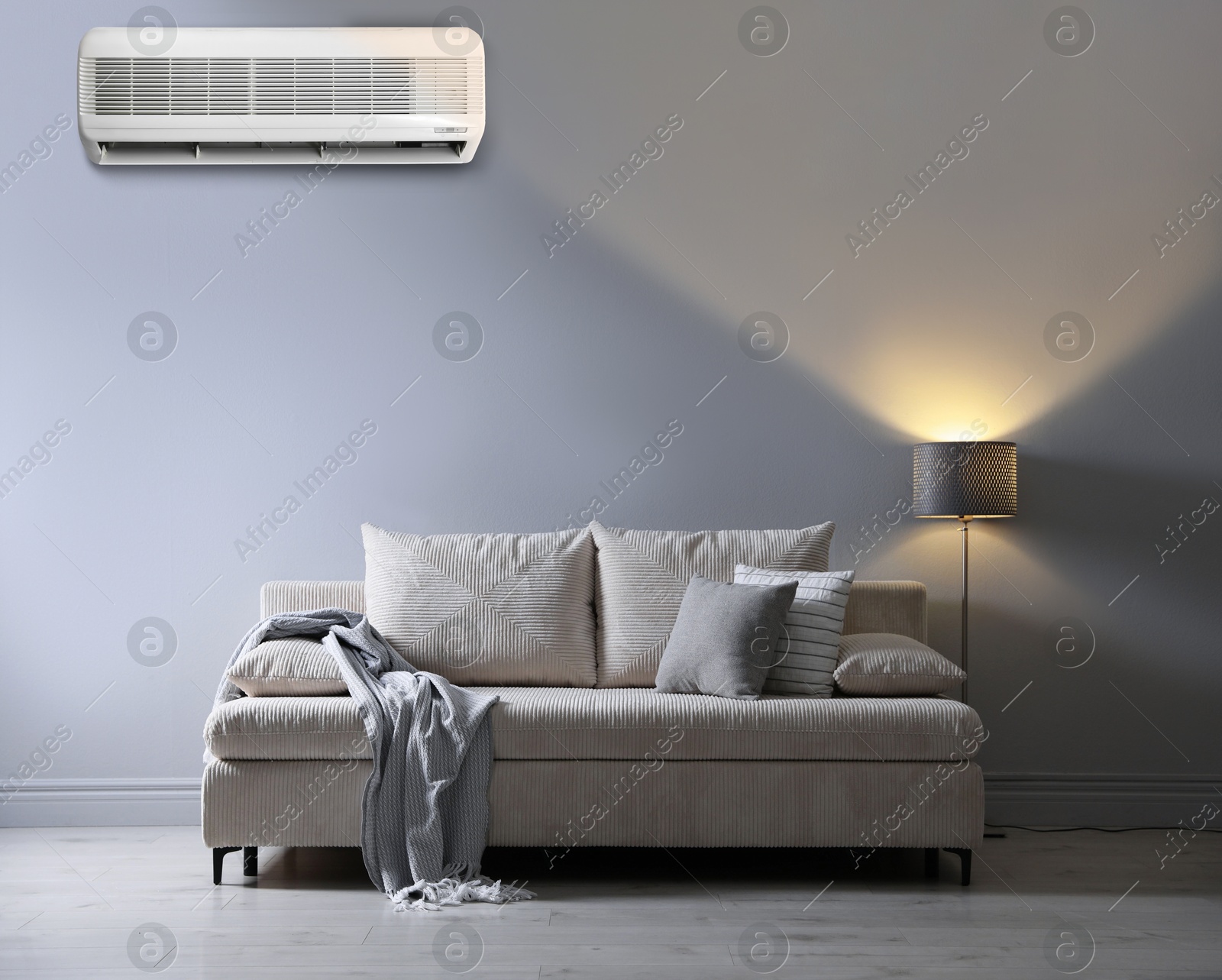 Image of Sofa with pillows, blanket and lamp near grey wall in room. Air conditioner keeping comfortable temperature indoors