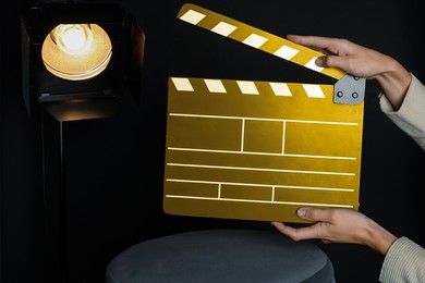 Image of Woman with golden clapperboard near spotlight on black background, closeup