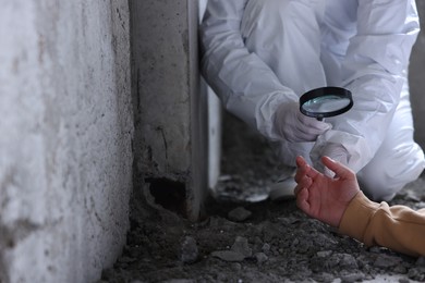 Photo of Forensic expert with magnifying glass working at crime scene with dead man's body inside abandoned building, closeup