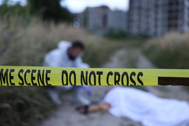 Photo of Forensic expert working at crime scene with dead body outdoors, focus on yellow police tape