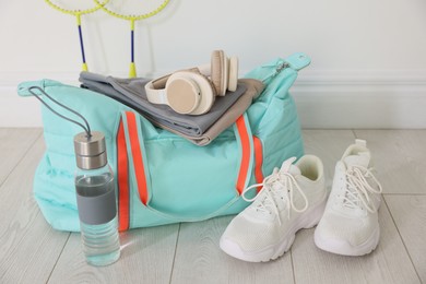Photo of Stylish sports bag with sportswear, sneakers, water bottle, headphones and badminton rackets on floor near white wall indoors